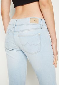 Jeans azul claro con cintura de media altura, patrón de olas sutil en el bolsillo trasero, parche de cuero con logotipo y detalles en hardware plateado.