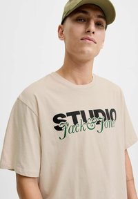 Beżowa bawełniana koszulka z krótkim rękawem, wyróżniająca się wyraźnym czarnym i zielonym napisem "STUDIO Jack & Jones". Luźny krój i gładka faktura.