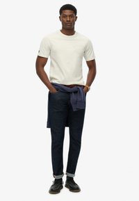 T-shirt gris clair à manches courtes avec logo texturé, pantalon en denim bleu foncé et chaussures noires à lacets. Sweat-shirt navy noué autour de la taille.