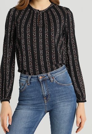 Kvinde iført en sort bluse med lange ærmer med lodrette blomsterstriber, stukket ned i højtaljede blå jeans.