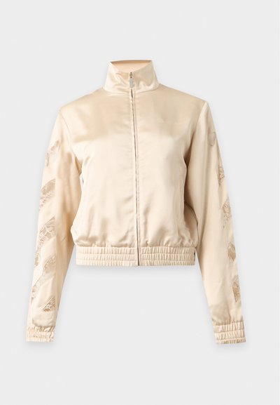Veste zippée en satin beige avec col haut, poignets et ourlet élastiques, présentant des motifs en dentelle découpée le long des deux manches.