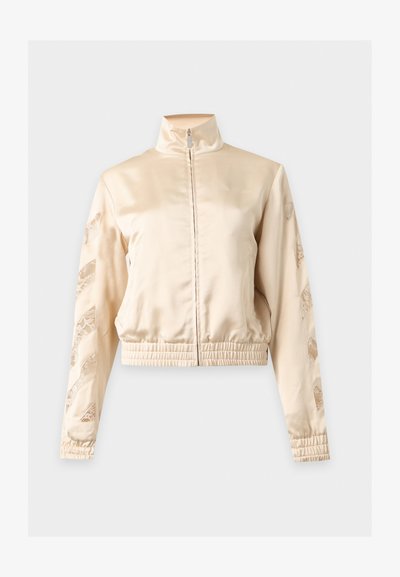 Veste zippée en satin beige avec col haut, poignets et ourlet élastiques, présentant des motifs en dentelle découpée le long des deux manches.