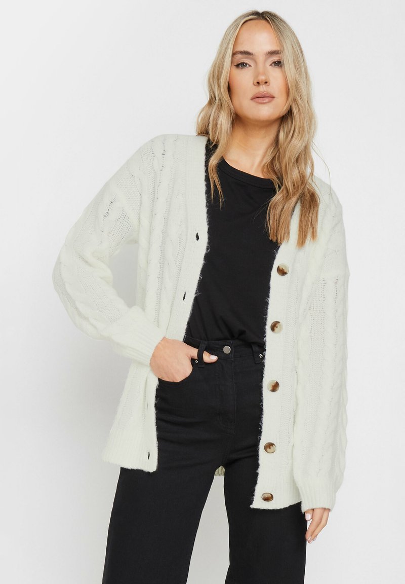 Cardigan verde chiaro a maglia cable con una vestibilità rilassata, caratterizzato da grandi bottoni marroni e polsini a coste, indossato sopra una maglietta nera e jeans.