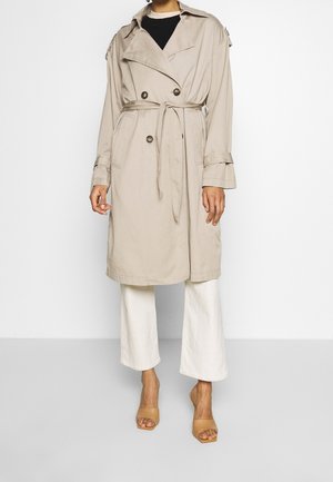 Persona che indossa un trench beige con cintura, pantaloni bianchi a gamba larga e tacchi aperti color cammello, in piedi su uno sfondo bianco semplice.