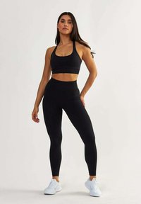 Un soutien-gorge de sport noir et un legging taille haute, tous deux en matériau lisse et extensible. Des chaussures de sport blanches complètent la tenue.