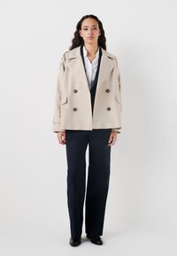 Trench coat beige doppiopetto con grandi bottoni, ampi rever e tasche laterali, abbinato a pantaloni navy a gamba larga e scarpe nere.