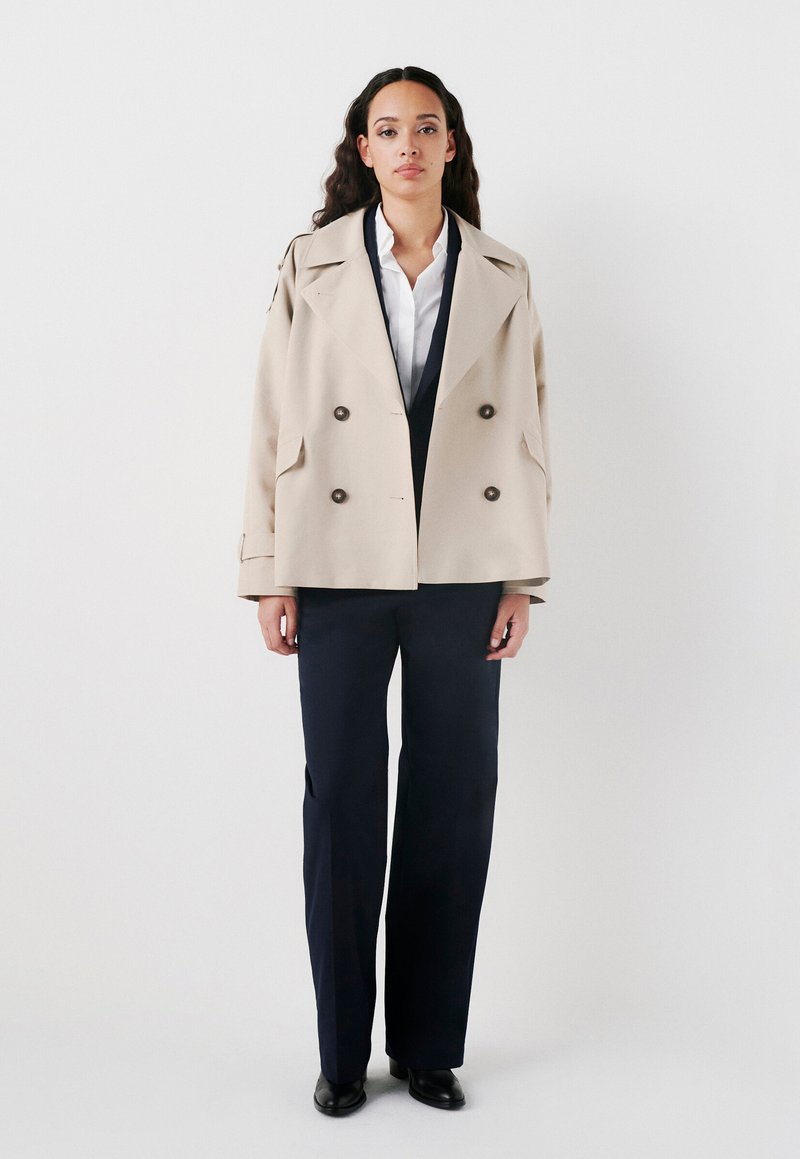 Trench coat beige doppiopetto con grandi bottoni, ampi rever e tasche laterali, abbinato a pantaloni navy a gamba larga e scarpe nere.