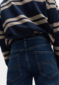 Jeans in denim blu scuro con un design aderente, caratterizzati da due tasche posteriori e un leggero effetto sbiadito. Abbinati a un top a righe blu navy e crema.