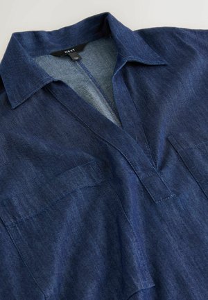 Cămașă din denim albastru închis cu guler, deschidere în formă de V și buzunare vizibile în față pe piept, așezată plat pe o suprafață deschisă la culoare.