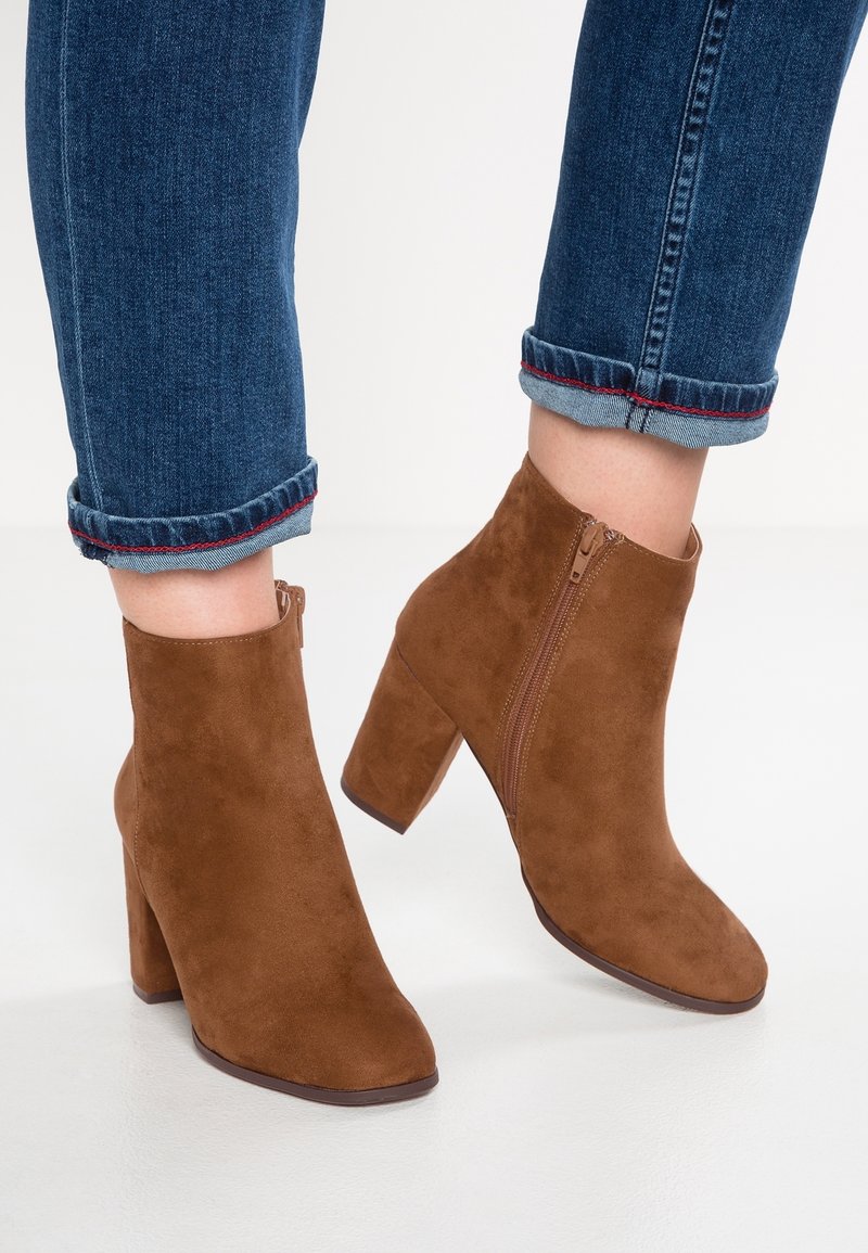 Personne portant un jean bleu retroussé et des bottines en daim marron à talons blocs avec des fermetures éclair latérales, debout sur une surface blanche.