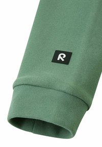 Manica di un felpa verde con una texture liscia; presenta un'etichetta quadrata nera con un logo bianco "R" sul polsino.