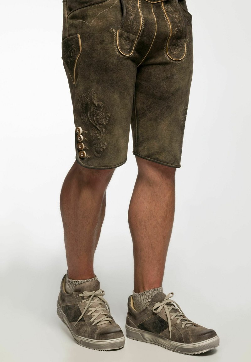 Braune Ledershorts mit aufwendiger Stickerei, seitlichen Taschen und Knopfdetails. Kombiniert mit lässigen grauen Sneakers und gestrickten Socken.