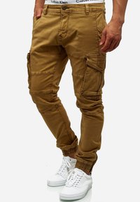 Pantalones cargo color caqui con puños elásticos, múltiples bolsillos laterales y detalles cosidos. Combinados con zapatillas blancas y una cinturilla visible.