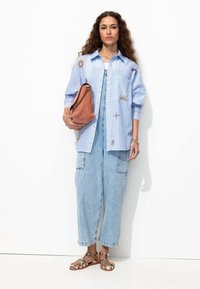 Chemise rayée bleu clair avec broderie de soleil et d'étoiles, salopette en denim, sandales plates dorées et sac en terre cuite décontracté avec accent chaîne.
