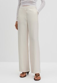 Beige wide-leg trousers in smooth fabric with a high waist, featuring side pockets and a tailored design. Paired with brown heels.