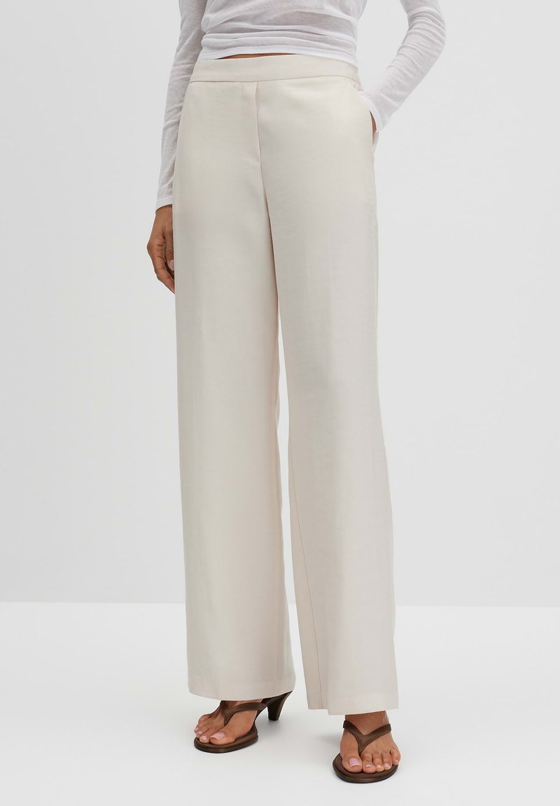 Beige wide-leg trousers in smooth fabric with a high waist, featuring side pockets and a tailored design. Paired with brown heels.