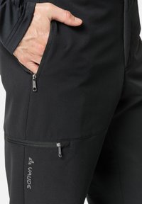 Pantaloni softshell neri con una texture liscia, dotati di due tasche con zip e un logo. Vestibilità slim con cuciture rinforzate per maggiore durata.