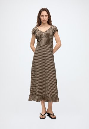 Vrouw draagt een bruin-beige geruite midi-jurk met korte pofmouwen en een gerimpelde zoom, staand met handen achter de rug.