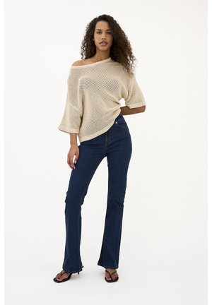 Frau steht mit der Hand in der Hüfte, trägt einen beigen schulterfreien Strickpullover, dunkelblaue Schlagjeans und schwarze Sandaletten mit Absatz vor weißem Hintergrund.