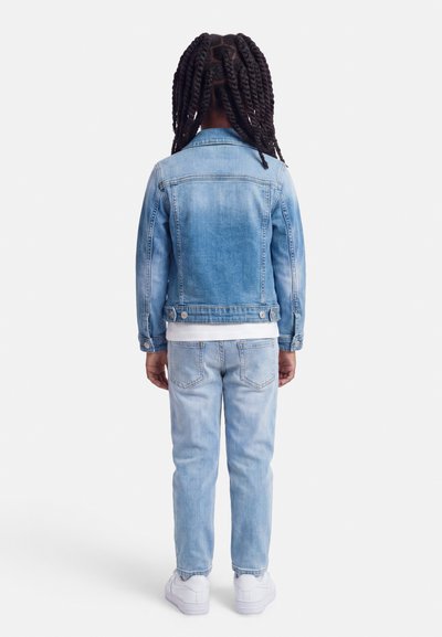 Παιδί με μακριά πλεξούδα στέκεται γυρισμένο προς τα πίσω, φοράει ανοιχτό μπλε τζιν μπουφάν και τζιν παντελόνι με λευκά αθλητικά παπούτσια σε λευκό φόντο.