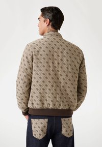 Veste bomber beige à motif avec un design en diamant et des bordures marron. Elle présente des poignets et une taille élastiques, associée à un pantalon en denim foncé.