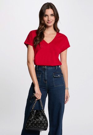 Camiseta roja de cuello en V con mangas cortas, que presenta detalles de encaje de ojales; combinada con pantalones de mezclilla oscuros de pierna ancha y cintura alta, y un bolso negro adornado.