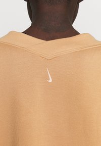 Sudadera de Nike en suave y ligero tejido marrón claro, con un diseño de cuello en V y un pequeño logo rosa claro en la parte trasera del cuello.