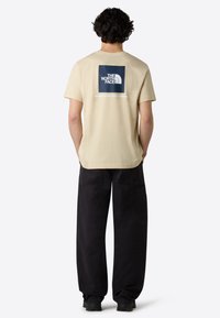 Beige tričko s krátkým rukávem s námořnickým čtvercovým grafickým zpracováním na zadní straně, obsahující nápisy "THE NORTH FACE" a "NEVER STOP EXPLORING".