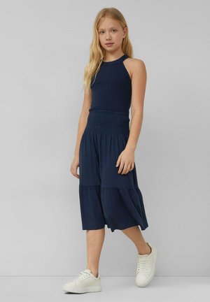 Junges Mädchen mit langen blonden Haaren, trägt ein navyfarbenes ärmelloses Top und einen passenden midilangen Rock, weiße Sneakers, geht vor einem grauen Hintergrund.