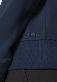 Sudadera azul oscuro con textura acanalada, cuello redondo y un discreto logo "DRYKORN" en el dobladillo. Confeccionada en una tela suave.