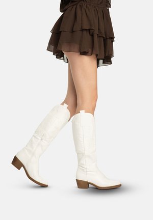 Ben iført lagdelt brun mininederdel og hvide knælange støvler med træblokhæle gående på ensfarvet baggrund.