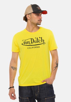 Homme portant un t-shirt jaune Von Dutch California, une casquette noire et beige, un jean foncé, avec des tatouages de papillon et d'araignée sur ses bras.