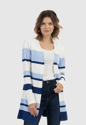 Frau trägt einen langen weißen Cardigan mit blauen horizontalen Streifen, weißes Top und dunkelblaue Jeans, steht mit einer Hand in der Tasche vor grauem Hintergrund.