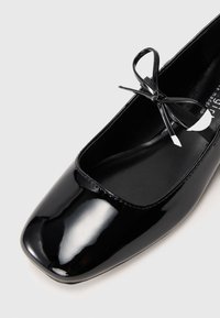 Bailarina em pato preto brilhante com ponta quadrada, apresentando um pequeno detalhe de laço na frente. Textura suave, design minimalista.