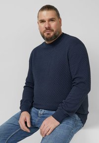 Granatowa bluza z teksturowanym wzorem diamentowym, ściągaczami na rękawach i okrągłym dekoltem, zestawiona z jasnoniebieskimi dżinsami. Modelka siedzi na tle szarego tła.