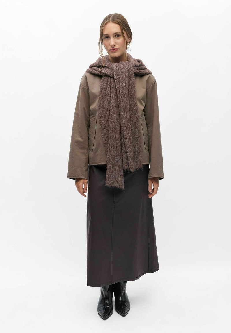 Femme vêtue d'une veste marron, d'une grosse écharpe marron, d'une longue jupe sombre et de bottes noires à bouts pointus, debout devant un fond blanc uni.
