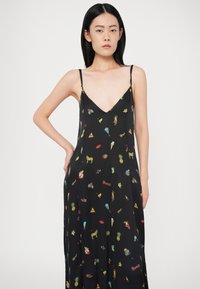 Frau mit langen schwarzen Haaren, die ein schwarzes Spaghettiträgerkleid mit bunten kleinen grafischen Drucken von Objekten und Tieren trägt.