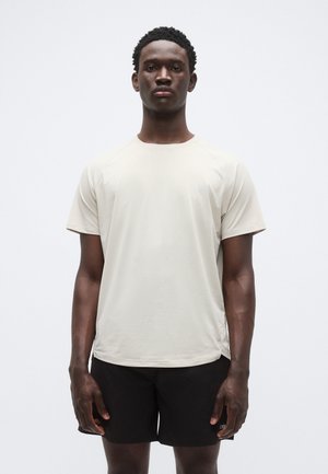 Gaiši bēšs sporta krekls ar apaļu kakla izgriezumu un īsām piedurknēm, ar gludu tekstūru un smalku logotipa detaļu, kopā ar melniem šortiem.