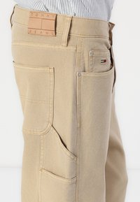 Beige Baumwolljeans mit mehreren Taschen und einem strukturierten Bund. Verfügt über ein Lederetikett mit dem Prägeaufschrift "TOMMY JEANS" und einen kleinen Flaggenakzent.