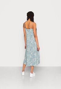 Mujer con pelo largo trenzado, vestida con un vestido midi sin mangas de estampado azul y blanco y zapatillas blancas, de pie de espaldas.