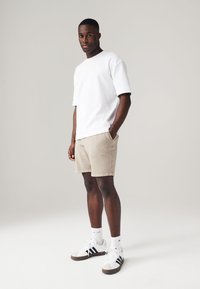 T-shirt bianca a maniche corte, pantaloni beige, sneakers bianche con strisce nere e calzini bianchi. Design semplice con vestibilità rilassata e dettagli minimi.