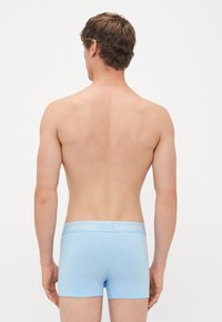 Hombre de pie con la espalda a la cámara, vistiendo calzoncillos boxer Lacoste de color azul claro, contra un fondo blanco liso.