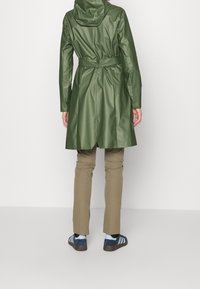 Zielony wodoodporny płaszcz z kapturem, przewiązany w talii, z plisami z tyłu, w połączeniu z beżowymi spodniami i niebieskimi trampkami z jasnoniebieskimi akcentami.