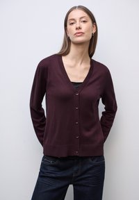 Cardigan bordeaux con scollo a V e maniche lunghe, bordo a coste e chiusura a cinque bottoni. Materiale in maglia leggera, abbinato a jeans in denim scuro.
