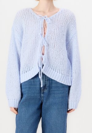 Kobieta nosząca jasnoniebieski dziergany kardigan związany trzema kokardkami na różowym topie i niebieskich dżinsach z wysokim stanem.