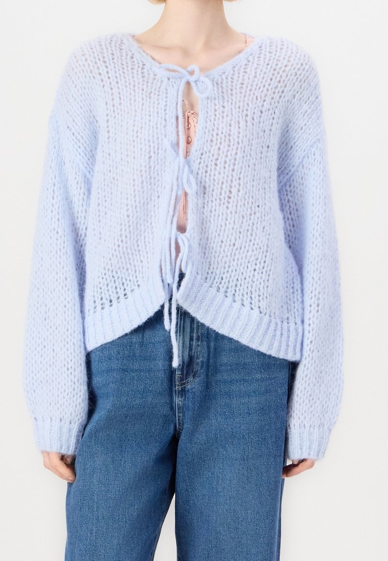 Femme portant un cardigan tricoté bleu clair noué avec trois nœuds sur un haut rose et un jean bleu taille haute.