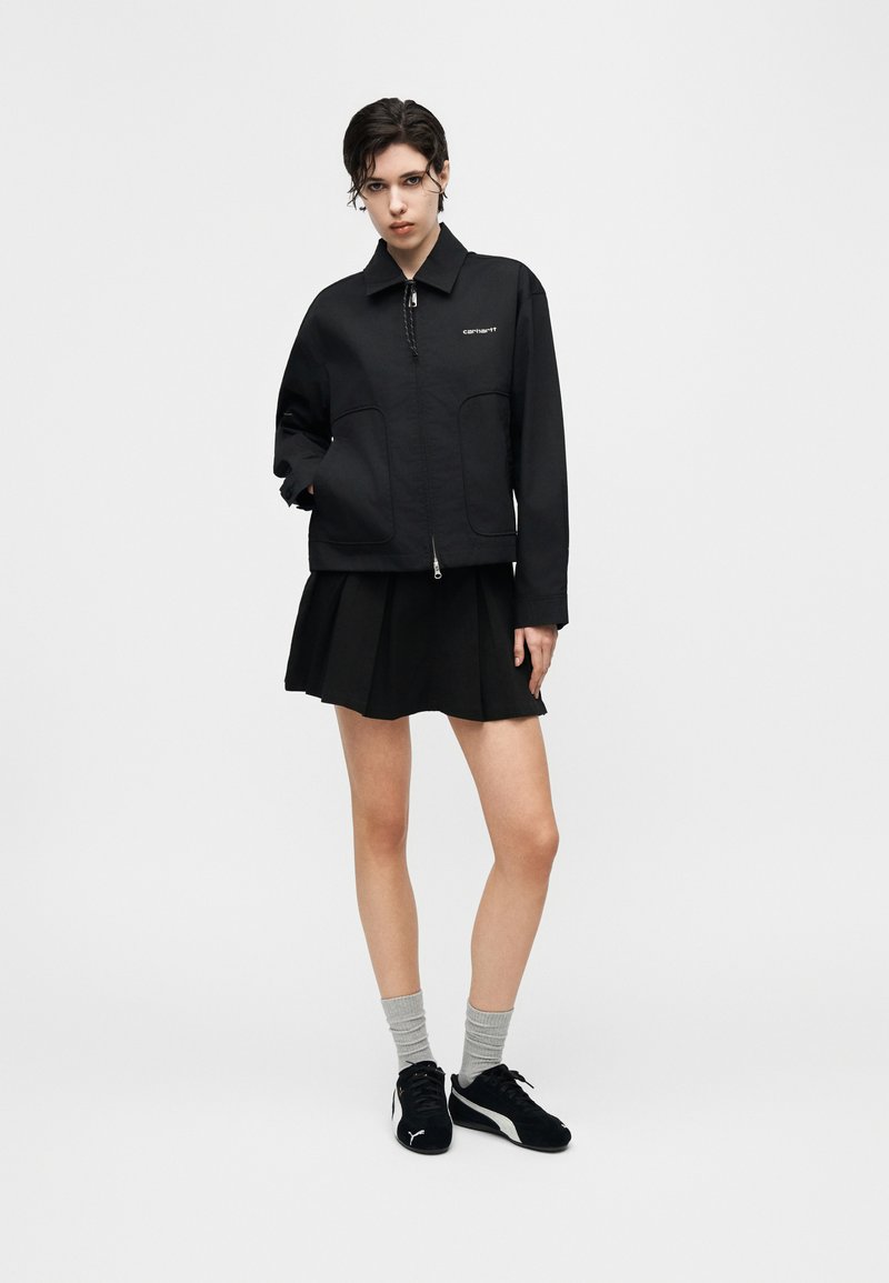 Giovane con capelli corti e scuri che indossa una giacca nera con cerniera, gonna nera a pieghe, calzini grigi e scarpe da ginnastica nere su sfondo bianco.