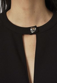 Schwarze Kleidung mit rundem Ausschnitt, die in der Mitte ein silbernes, rundes Verschlussdetail aufweist. Glatte Textur und minimalistisches Design.