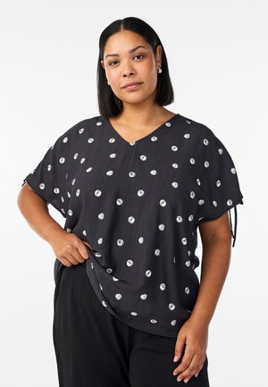 Kvinde med glat sort hår iført sort løstsiddende top med hvide polkaprikker og sorte bukser, poserer med den ene hånd i lommen.