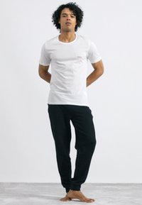 Un t-shirt en coton blanc avec un petit logo rouge "HUGO", associé à un pantalon noir décontracté, se tenant sur une surface gris clair.